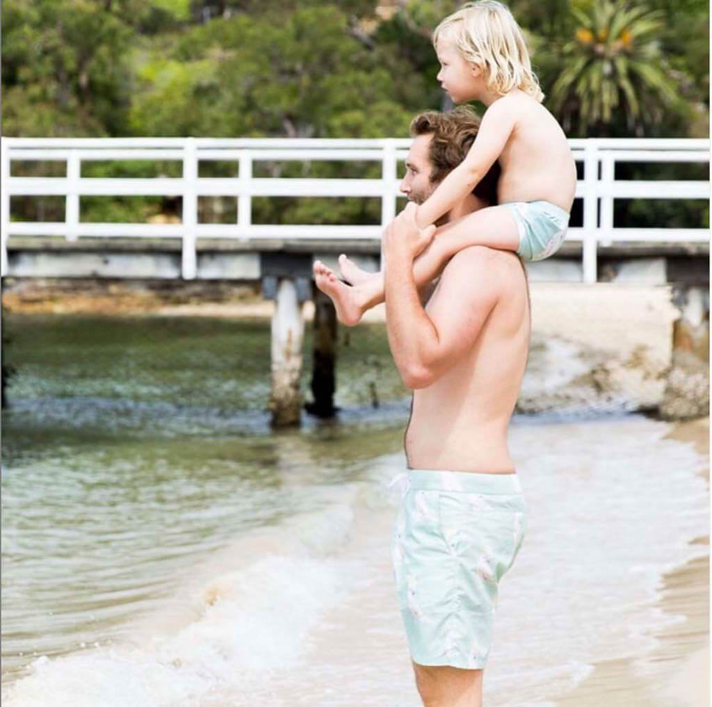 Man Wearing Green Cockatoo Men's Boardshorts While At Beach With Boy On His Shoulders