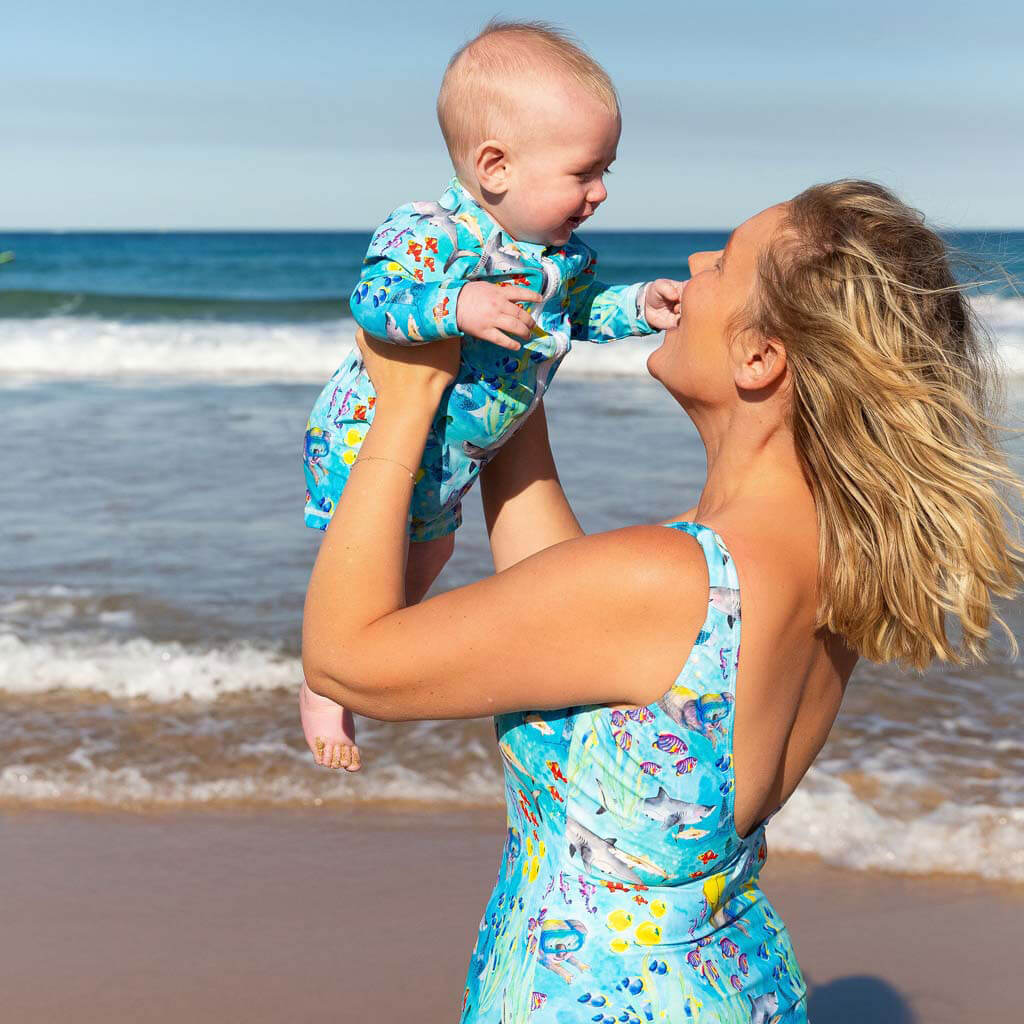 Woman on Beach Holding Baby Up Wearing Great Barrier Reef Women's One Piece Sleeveless Swimsuit. Baby In Matching Swimmers.