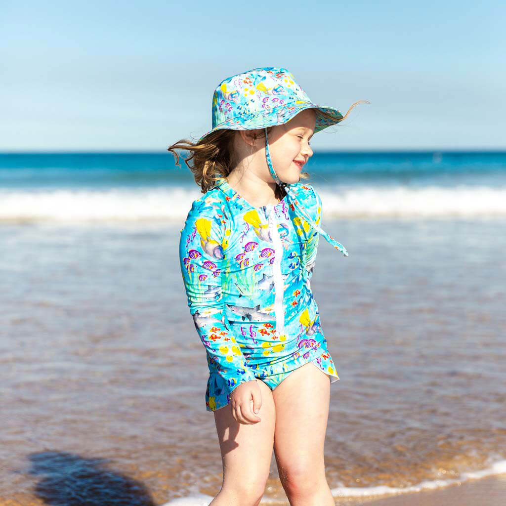 Smiling girl posing on beach wearing a Cheeky Chickadee Great Barrier Reef print swimsuit and beach hat