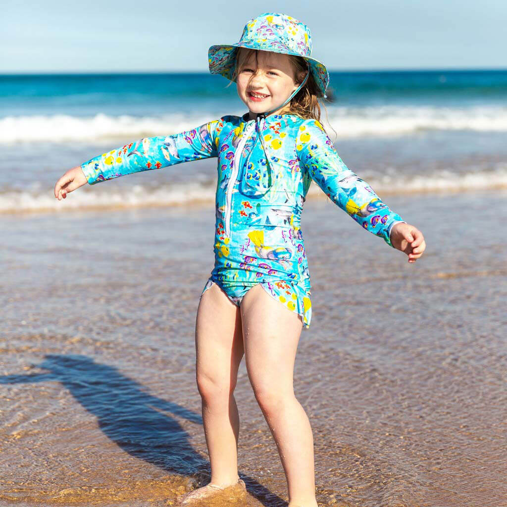 Smiling girl standing on beach wearing a Cheeky Chickadee Great Barrier Reef print swimsuit and beach hat
